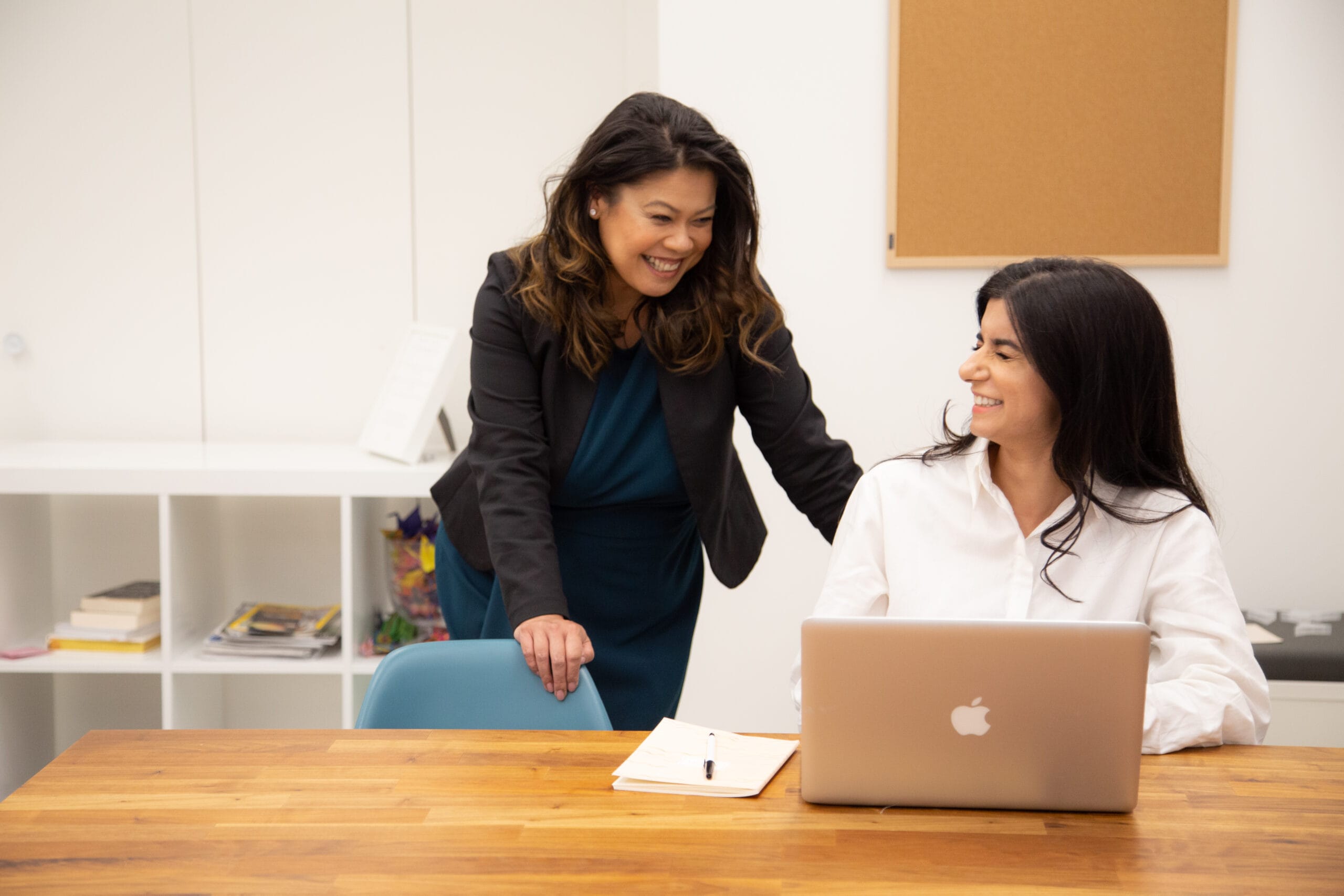 Woman boss checking in on woman employee working on her laptop, both laughing