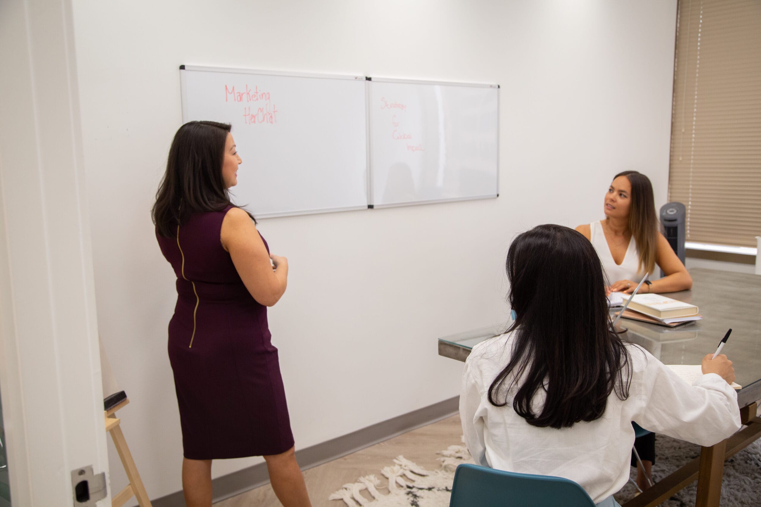 Woman leading meeting and writing on whiteboard, two other women listen closely